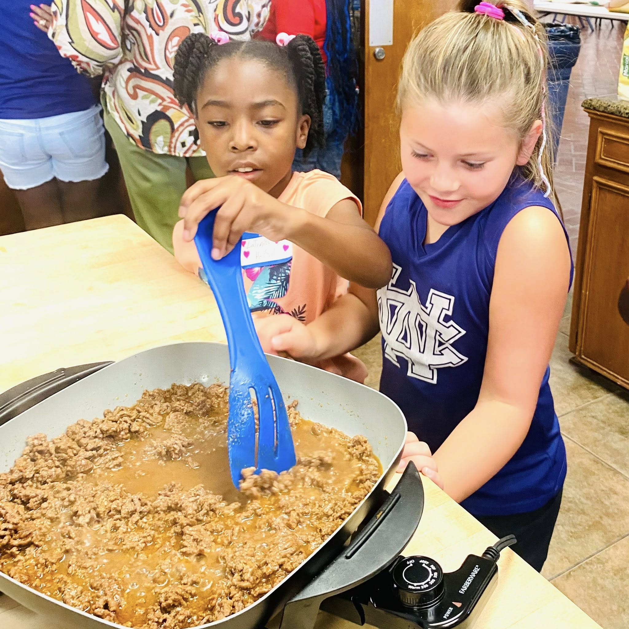 One girl watching another girl stir meat in an electric skillet.